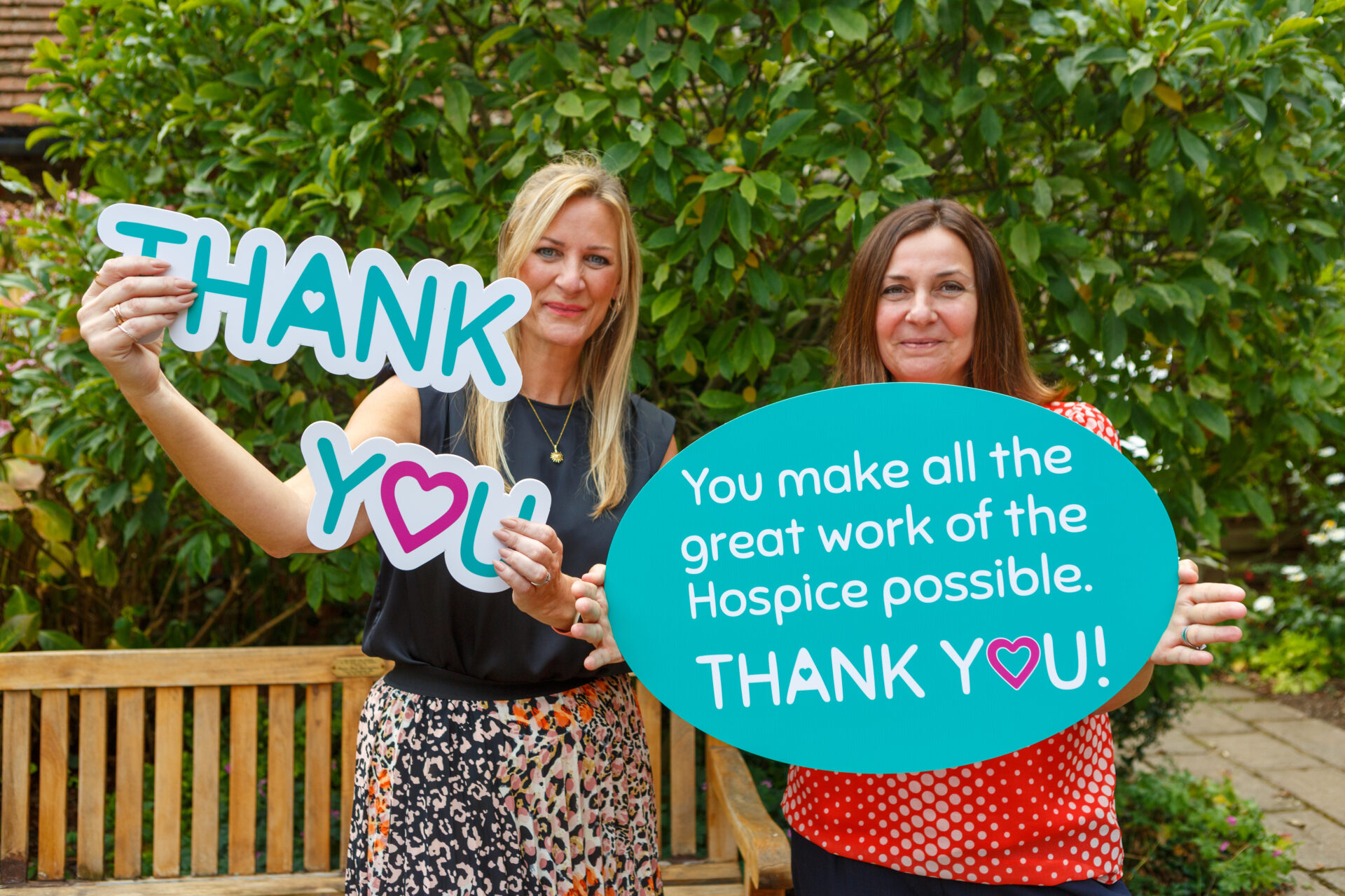 Two women holding up signs which say 'you make all the great work of the Hospice possible. Thank You'. In the Hospice garden