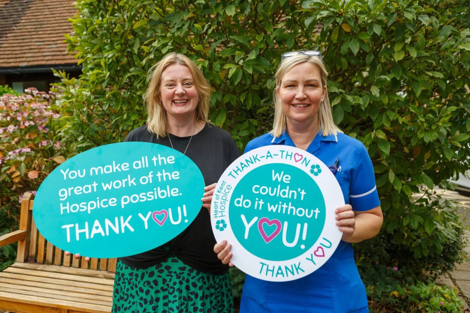 Two ladies standing in the hospice garden holding 'thank-a-thon' signs