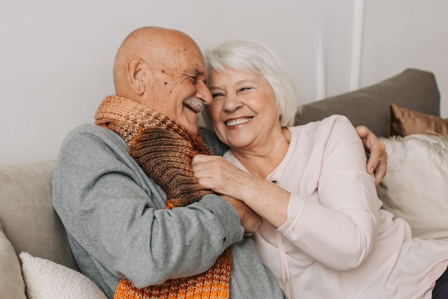 Man in Gray Sweater Hugging a woman in a pink cardigan, both smiling looking at each other, sat on a sofa.